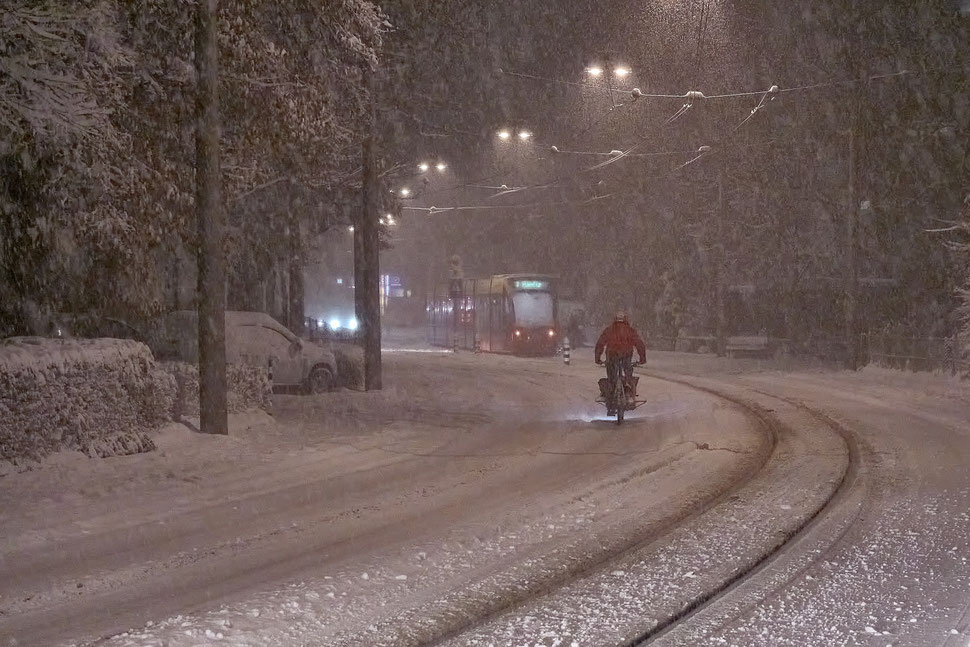 The image shows a night photograph of a man on a bicycle riding a snow covered street during a snowstorm. 