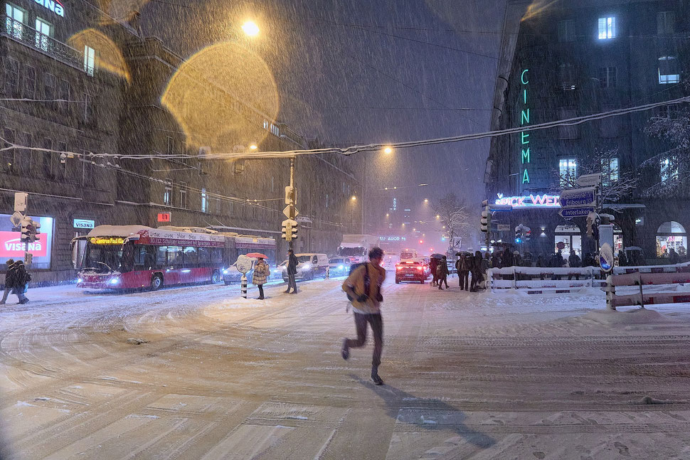 The image shows a nocturnal photograph of a snow covered street in a busy city with people and cars and a bus during a winter storm. 