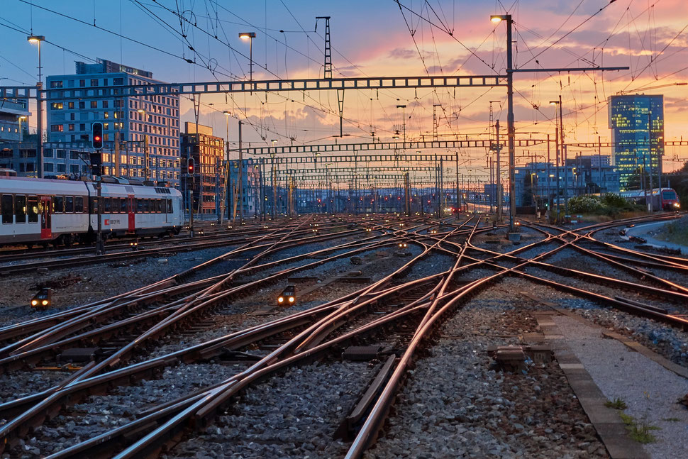 The image shows a photograph of main train tracks, building and train compositions at Zurich Main Station while the sun is setting.