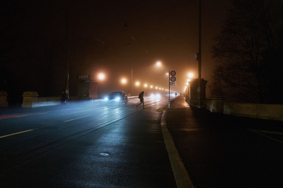 The image shows the photograph of approaching cars, headlights, streetlights and a woman crossing a street on a foggy night. 