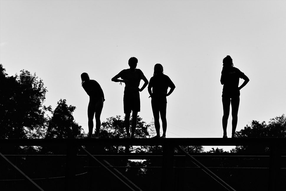The image shows the photograph of four young people standing on the railing of a bridge waiting to jump into the water. The location is Wohlensee near Bern.