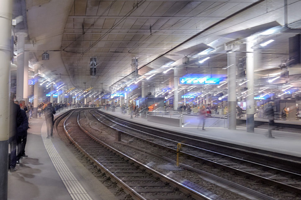 The image shows the blurry photograph of train tracks and people moving and standing on a platform of Bern main station.