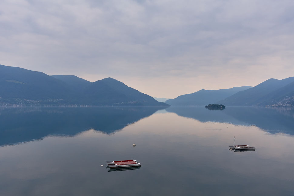 The image shows the photograph of a part of Lake Maggiore with one of the Brissago islands and the shore with mountains. The water is very still and there are two bathing rafts in the foreground.