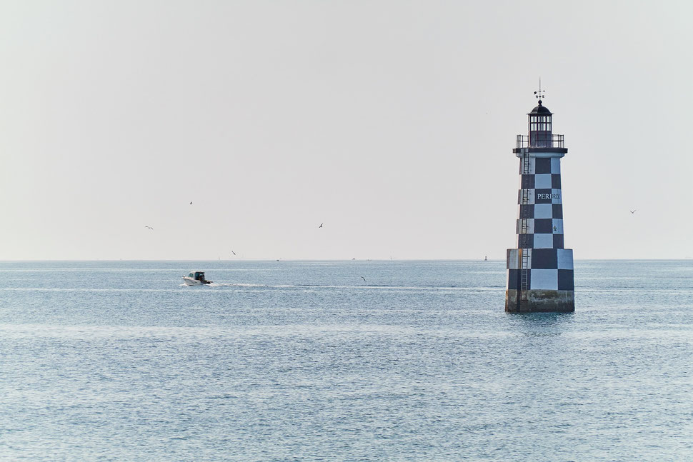 The image shows the photograph of a lighthouse with some birds in the air and a boat moving away. The location is Loctudy on the coast of Brittany.