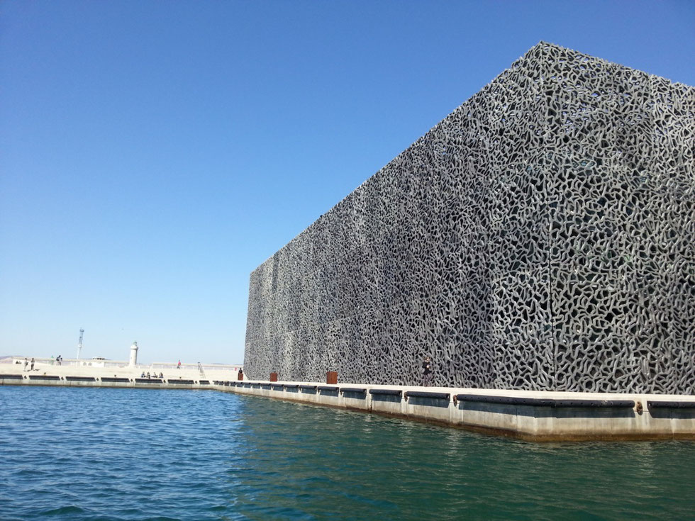 Le mucem est un grand rectangle couvert de son voile ajouré de béton gris, à l'entrée du vieux-port de Marseille, relié par le haut au fort Saint-Jean. #Provence #Art #AyniArt