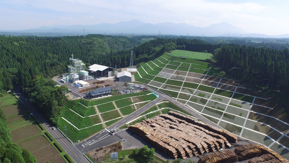 発電所全景（循環流動層ボイラ・蒸気タービン・冷却設備・徐鉄装置・チップヤード・破砕設備・管理棟・貯木場等）背景は霧島連山