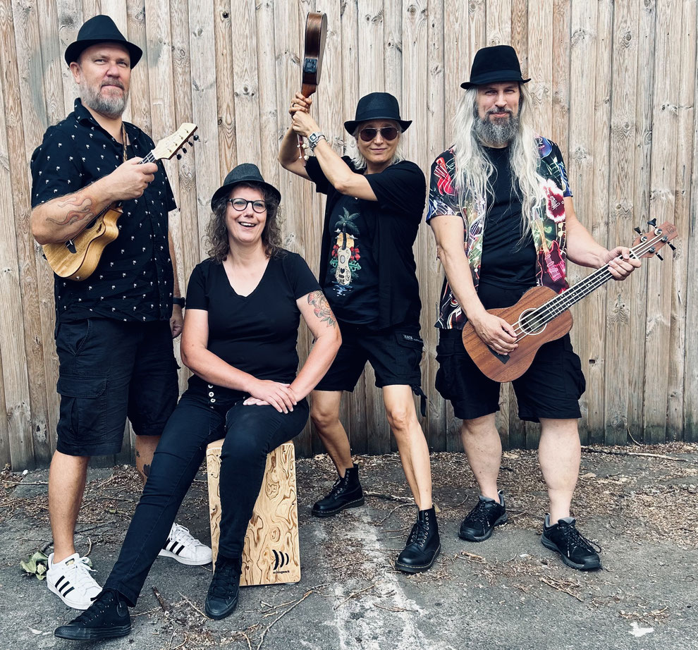 Drei Männer und eine Frau mittleren Alters in dunkler, lässiger Kleidung posieren für das Bandfoto.