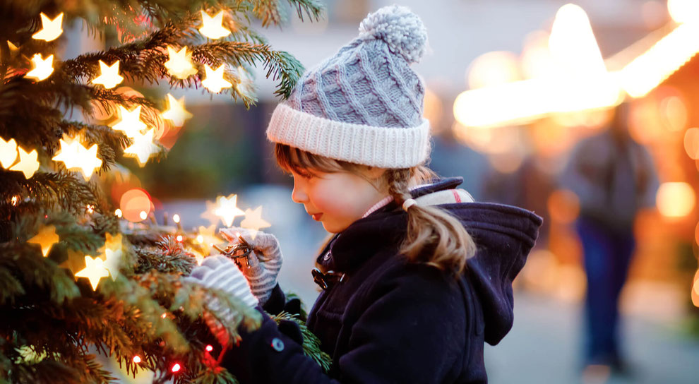 Fröhliches Mädchen im Schnee am traditionellen Weihnachtsmarkt, steht lächelnd neben einem festlich beleuchteten Tannenbaum.
