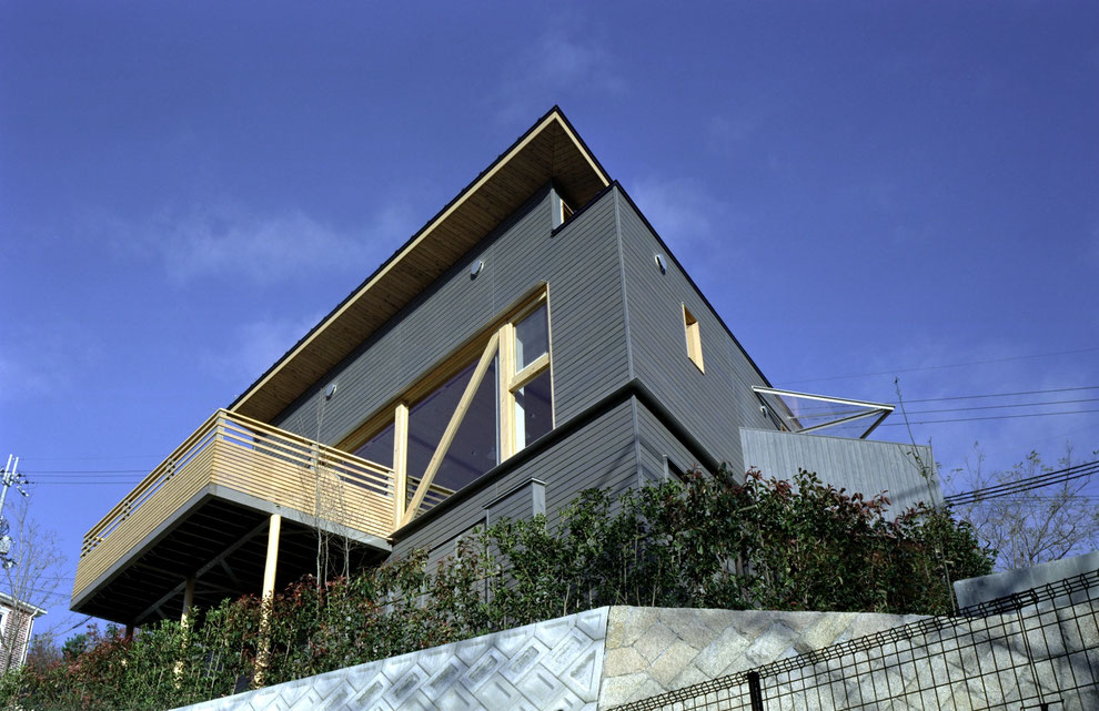 平賀敬一郎建築研究所が設計した、斜面地に建つモダンな木造住宅の外観写真。深い軒とシャープな片流れ屋根、木製の格子バルコニーが特徴的な、空に映えるスタイリッシュなデザイン。