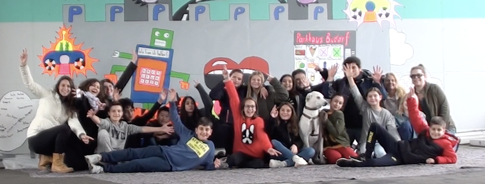 Klassenfoto vor einer bunt bemalten Wand. Die Schülerinnen und Schüler sitzen, liegen oder hocken, strecken die Arme in die Luft und lachen.