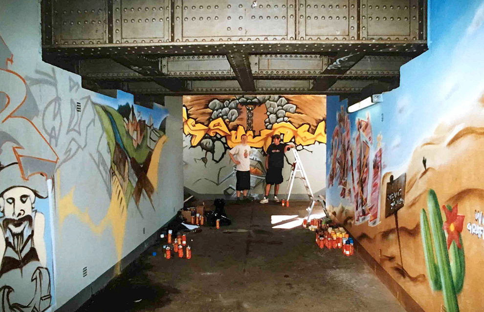 zwei junge Männer sprayen einen Graffiti Auftrag in der Unterführung eines Bahnhofs