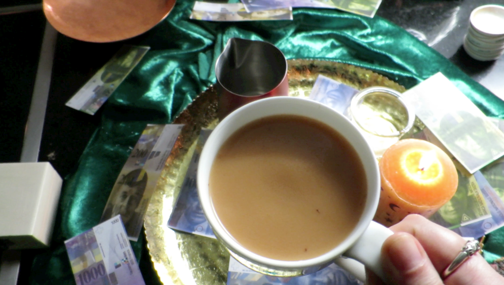 Eine Nahaufnahme einer Person mit einer Kaffeetasse vor einem esoterischen Tisch mit Papiergeld und Kerzen, im Hintergrund ein grünes Samttuch.
