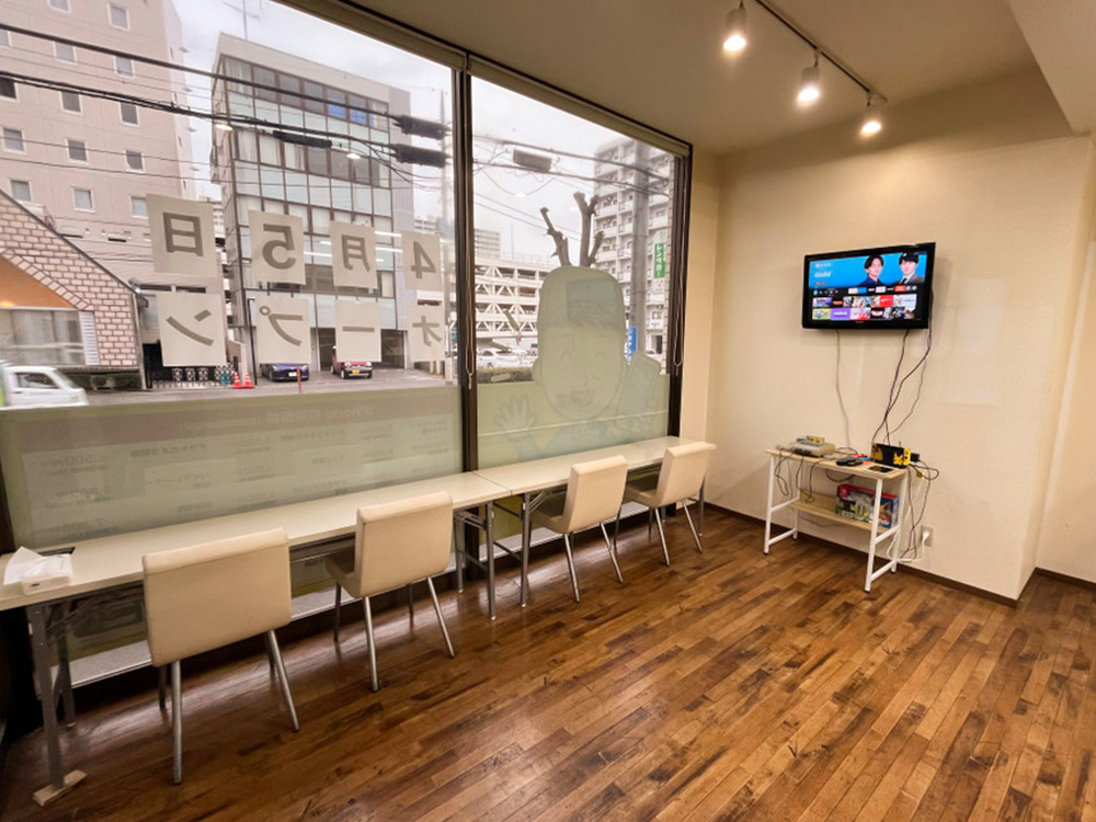 リペア土浦店の店内に、窓際のカウンター席と椅子、壁掛けテレビが設置されている写真。修理待ちの時間を過ごせる待機スペースの様子。