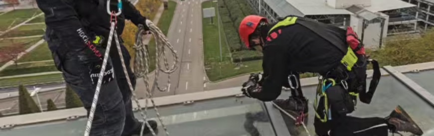 2 Personen in Persönlicher Schutzausrüstung gegen Absturz arbeiten an einer Dachkante und sind gegen die Absturzgefahr mit einem mitlaufenden Auffanggerät gesichert