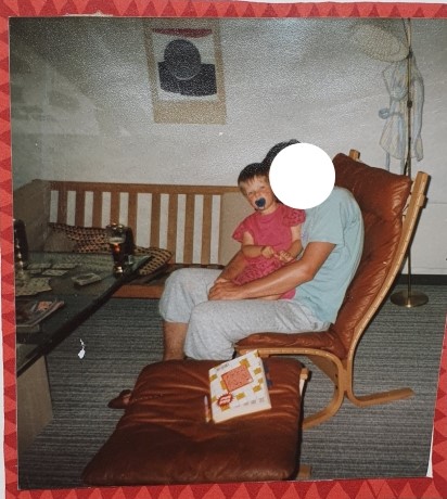Auf dem Urlaubsbild von 1992 bin ich knapp 2 Jahre alt. Mein Papa sitzt in einem braunen Sessel. Mit blauem Schnuller im Mund sitze ich auf seinem Schoß und er umarmt mich. Ich trage ein pinkes Oberteil und eine pinke Hose.