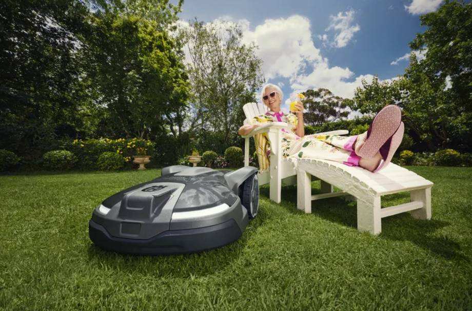 Eine ältere Dame entspannt sich mit einem Drink in der Hand auf einem Liegestuhl, während ihr Husqvarna Automower, ein zuverlässiger Mähroboter, den Rasen in einem sonnigen Garten bearbeitet.