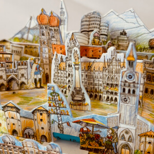 Zwei Fotos eines dreidimensionalen Modells von München. Alle Teile sind gezeichnet, auf dünne Pappe gedruckt und ausgestanzt. Sie werden wie ein dreidimensionales Puzzle zusammengesteckt. Die Teile passen alle in eine Streichholzschachtel. Die kleine Schachtel eignet sich wunderbar als Geschenk für Reiselustige oder als Erinnerung.