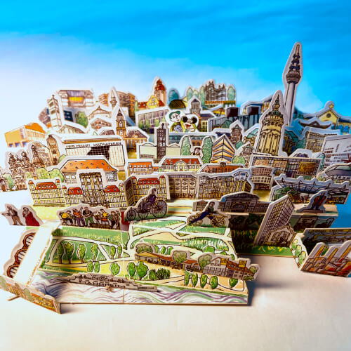 Zwei Fotos eines dreidimensionalen Modells von Mannheim. Alle Teile sind gezeichnet, auf dünne Pappe gedruckt und ausgestanzt. Sie werden wie ein dreidimensionales Puzzle zusammengesteckt. Die Teile passen alle in eine Streichholzschachtel. Die kleine Schachtel eignet sich wunderbar als Geschenk für Reiselustige oder als Erinnerung.