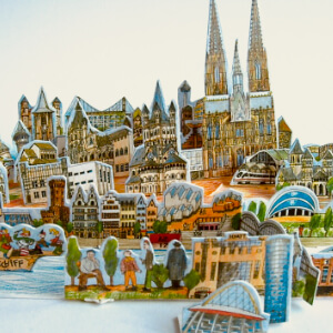 Zwei Fotos eines dreidimensionalen Modells von Köln. Alle Teile sind gezeichnet, auf dünne Pappe gedruckt und ausgestanzt. Sie werden wie ein dreidimensionales Puzzle zusammengesteckt. Die Teile passen alle in eine Streichholzschachtel. Die kleine Schachtel eignet sich wunderbar als Geschenk für Reiselustige oder als Erinnerung.