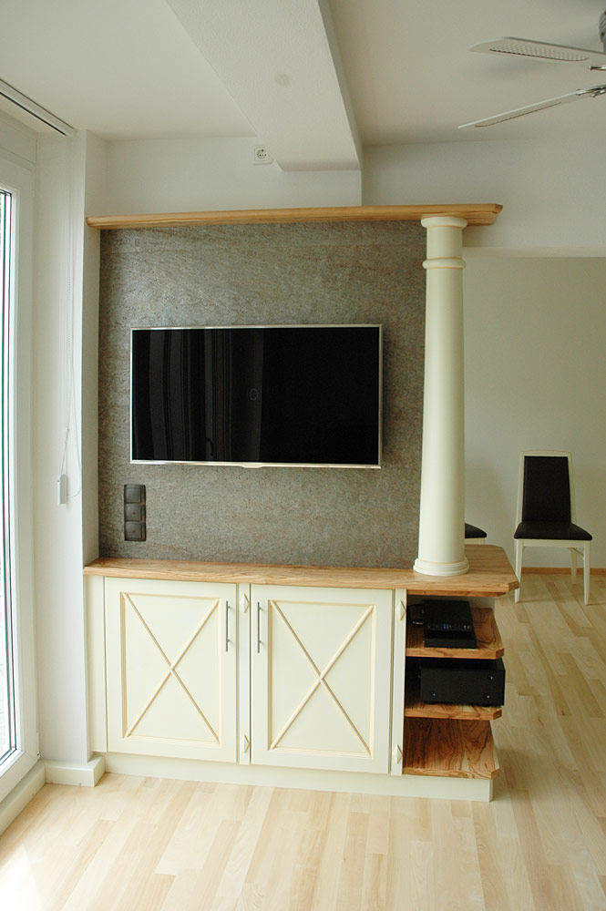 Eine besondere Fernsehwand: Olivenholz mit weiß lackierten und beige patinierten Fronten. Die Steinwand hinter dem Fernseher ist tatsächlich aus Stein - einer dünnen Schicht aus echtem Stein, die wir selbst verarbeiten und die den Raum beruhigt.
