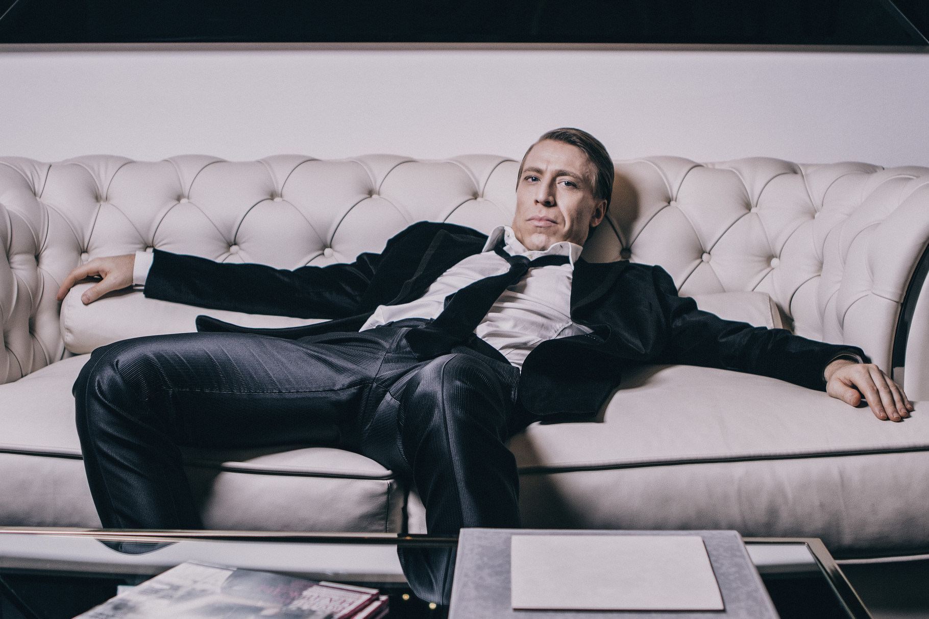 Ottaviano Blitch, Italian actor and performer, elegant editorial portrait reclining on a tufted sofa in a dark suit, attore performer italiano ritratto elegante editoriale