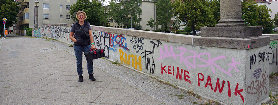 Ruth Kleinöder steht auf einer Brücke und hält einen Werkzeugkoffer in der Hand. Die Brückenmauer ist mit Graffitis besprüht.