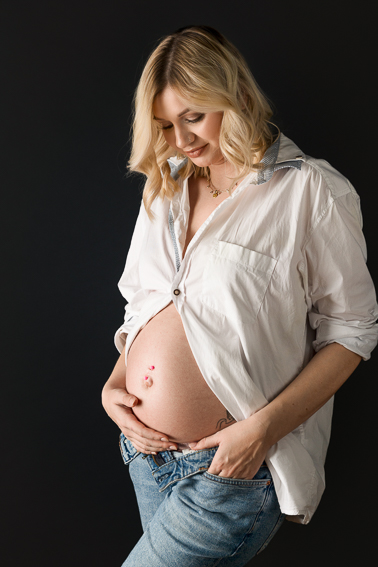 Schwangere Frau im Fotostudio, steht entspannt vor dunklem Hintergrund und hält den Babybauch