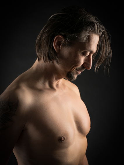 Männliches Boudoir-Portrait im Studio mit seitlicher Lichtführung, Profilansicht eines Mannes mit nacktem Oberkörper vor dunklem Hintergrund