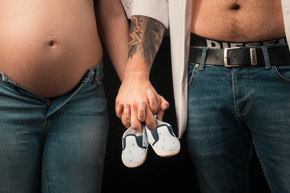 Paar im Studio steht nebeneinander und hält kleine Babyschuhe vor dem Babybauch