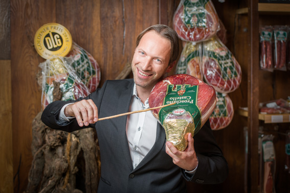 Inszenierte Werbefotografie eines Unternehmers mit verpacktem Schinkenprodukt in einer Fleischerei, Produktpräsentation im Verkaufsumfeld