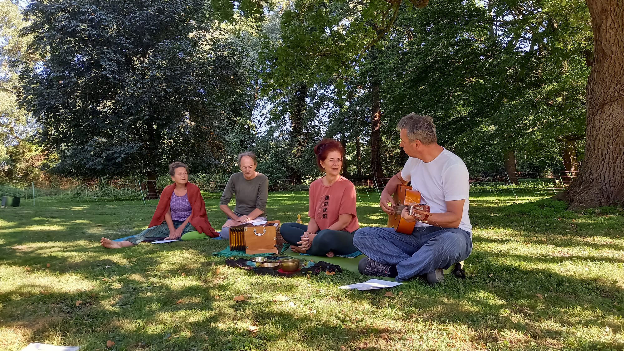 Vier Menschen sitzen auf einer Wiese unter einen Baum, ein Mann spielt Gitarre 