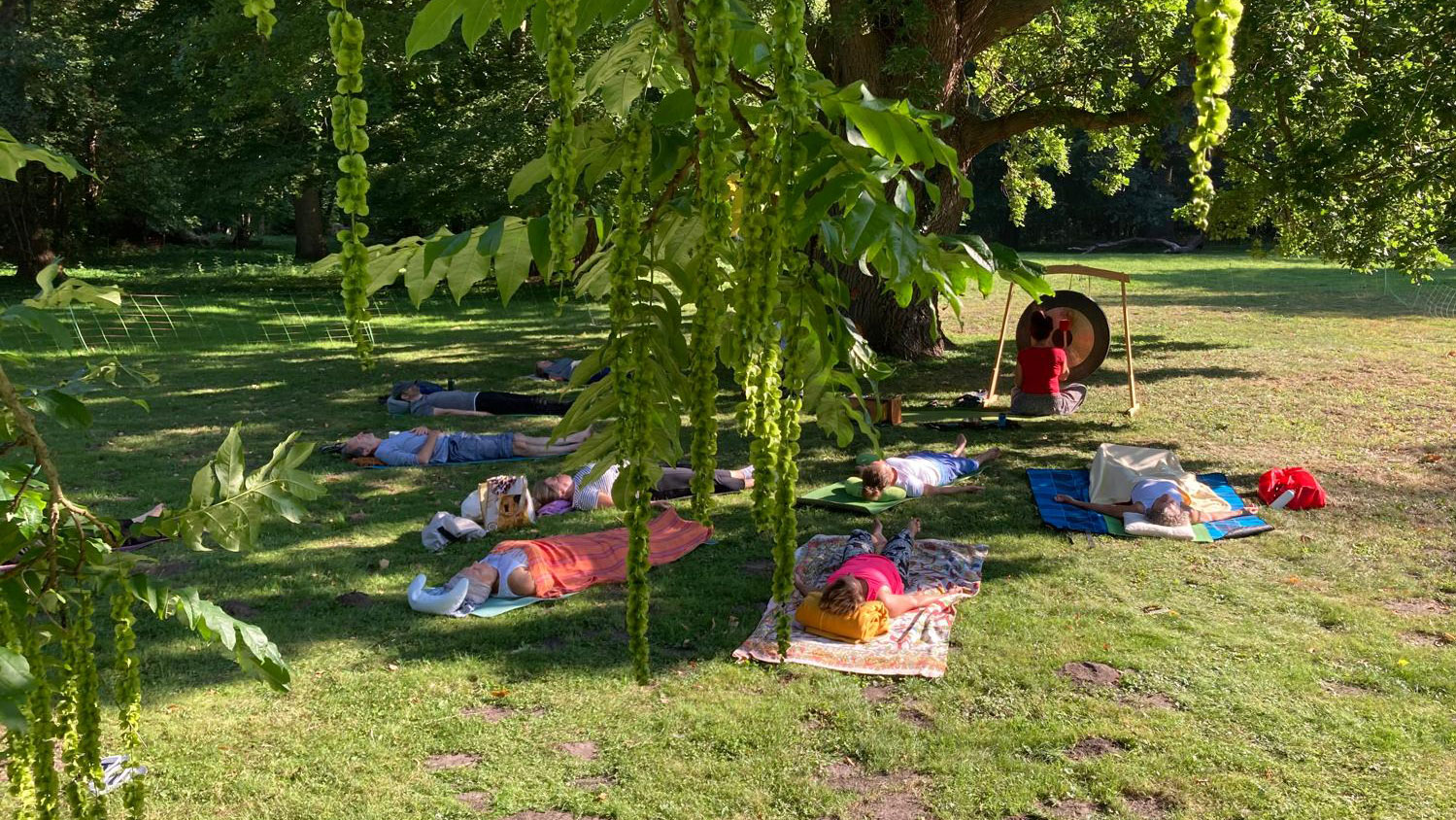 Mehrere Menschen liegen auf Yogamatten auf der Wiese, im Hintergrund schlägt eine Frau auf einen Gong 