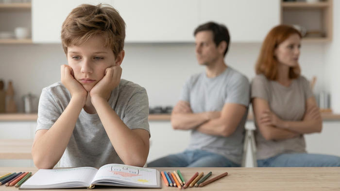 Un bambino dall’espressione triste e preoccupata siede al tavolo con un quaderno e delle matite colorate; sullo sfondo, sfocati e distanti, i genitori mostrano la tensione di una crisi di coppia che può avere un impatto sui figli.