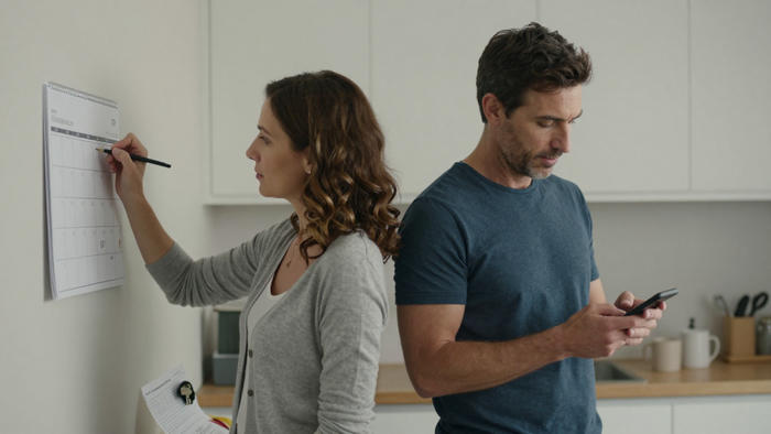 Una donna segna un calendario mentre un uomo guarda il cellulare in cucina: una scena di vita quotidiana che esprime distanza e gestione pratica nella coppia.