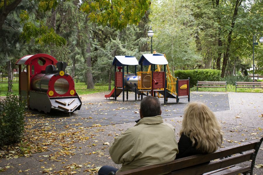 Ein Mann und eine Frau sitzen auf einer Parkbank und schauen auf einen leeren Kinderspielplatz. (Symbolbild: DianaZG)