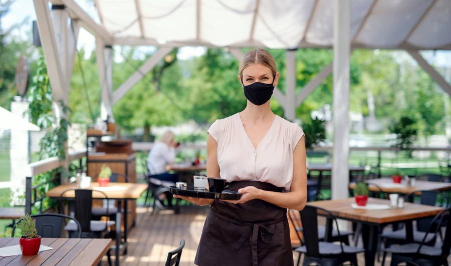 Eine Kellnerin mit Maske serviert auf der Terrasse eines Restaurants (Symbolbild: iStock/Halfpoint)