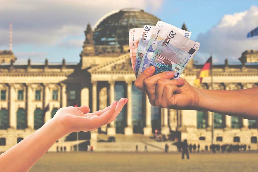 Eine Hand reicht einer anderen Hand Geldscheine. Im Hintergrund ist der Reichstag zu sehen. (Symbolbild: Ralph)