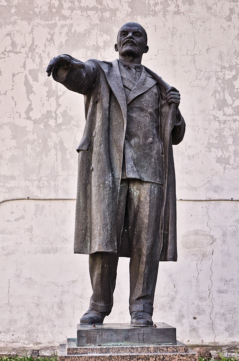Bronze statue of a man in a coat with one arm outstretched, standing on a stone pedestal against a weathered wall.