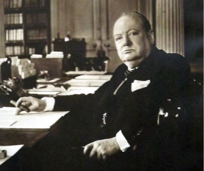 A black-and-white photo of Winston Churchill seated at a desk, holding a cigar, with a serious expression in a formal office setting.