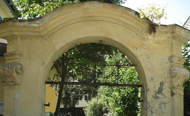 Der (zu renovierende) barocke Torbogen mit geschweiftem Giebel und schmiedeeisernem Laub- und Bandelwerk