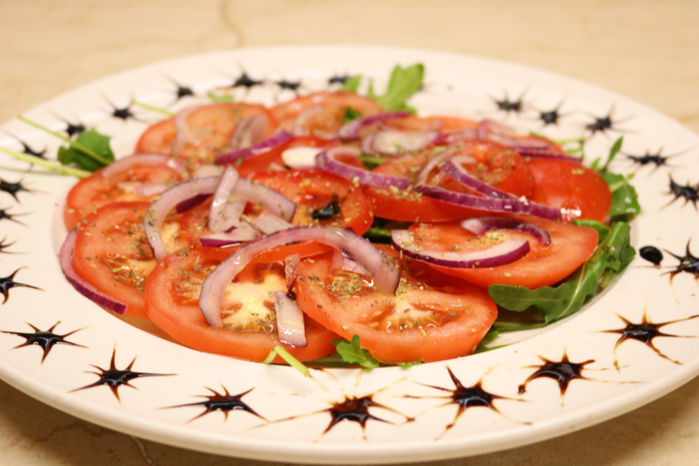 Tomatensalat mit roten Zwiebeln, Olivenöl, Oregano und Crema di Balsamico, auf einem Rucolabett