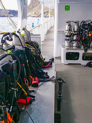 Photo of many oxygen tanks, BCDs, and weight belts on the deck of a dive boat
