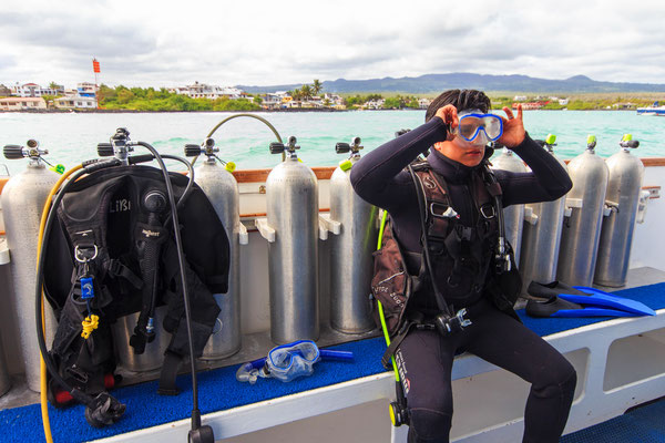 A diver adjusts their goggles and is in a wet suit and BCD and sits in a dive boat