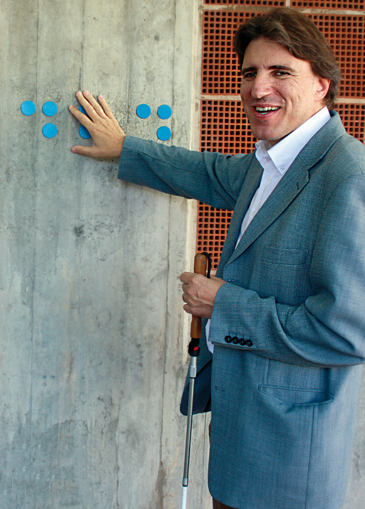 Manuele Bertoli touches a colored Braille composition on a concrete wall, holding a white cane