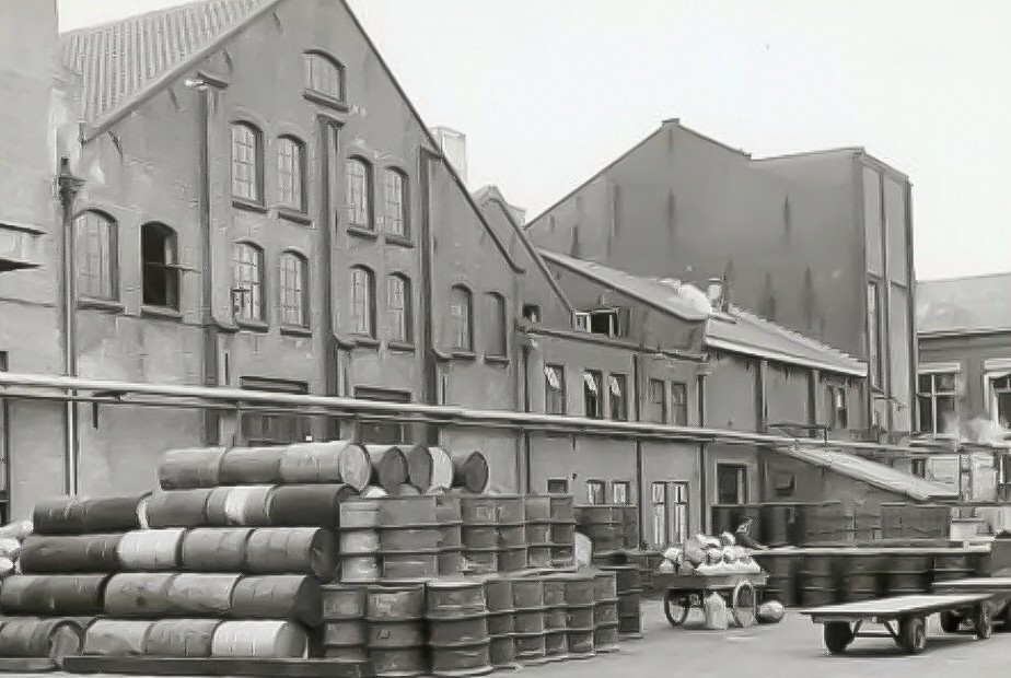 Oude fabrieksgebouwen van Calvé aan de Wateringseweg, waar de nieuwe levensmiddelenfabriek gebouwd zal worden. Op de voorgrond liggen stapels olievaten en rechts staat een lorry met links daarvan een bakfiets met olievaten, 1964