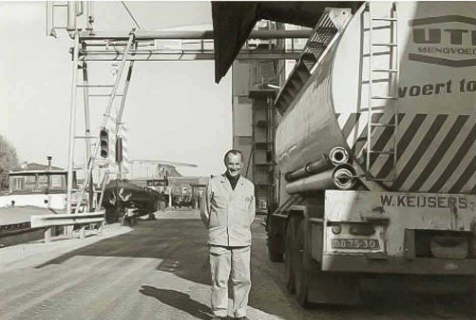P. Stigter naast een tankwagen op het terrein van de Calvéfabrieken aan de Wateringseweg. Op de achtergrond de Kolenhavenbrug met links schepen in de haven gezien naar het oosten. 1975
