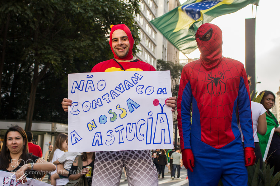 protesters wearing funny superhero costumes holding a sign and a Brazilian flag in background