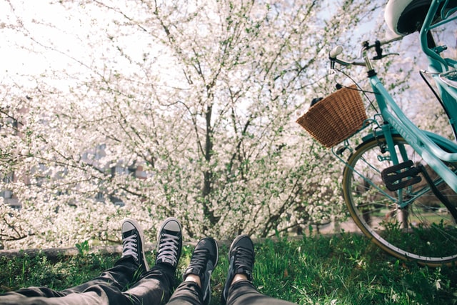 four feet in sneakers on two people lying on the green grass facing a cherry tree in full blossom and a light blue bike with a basket