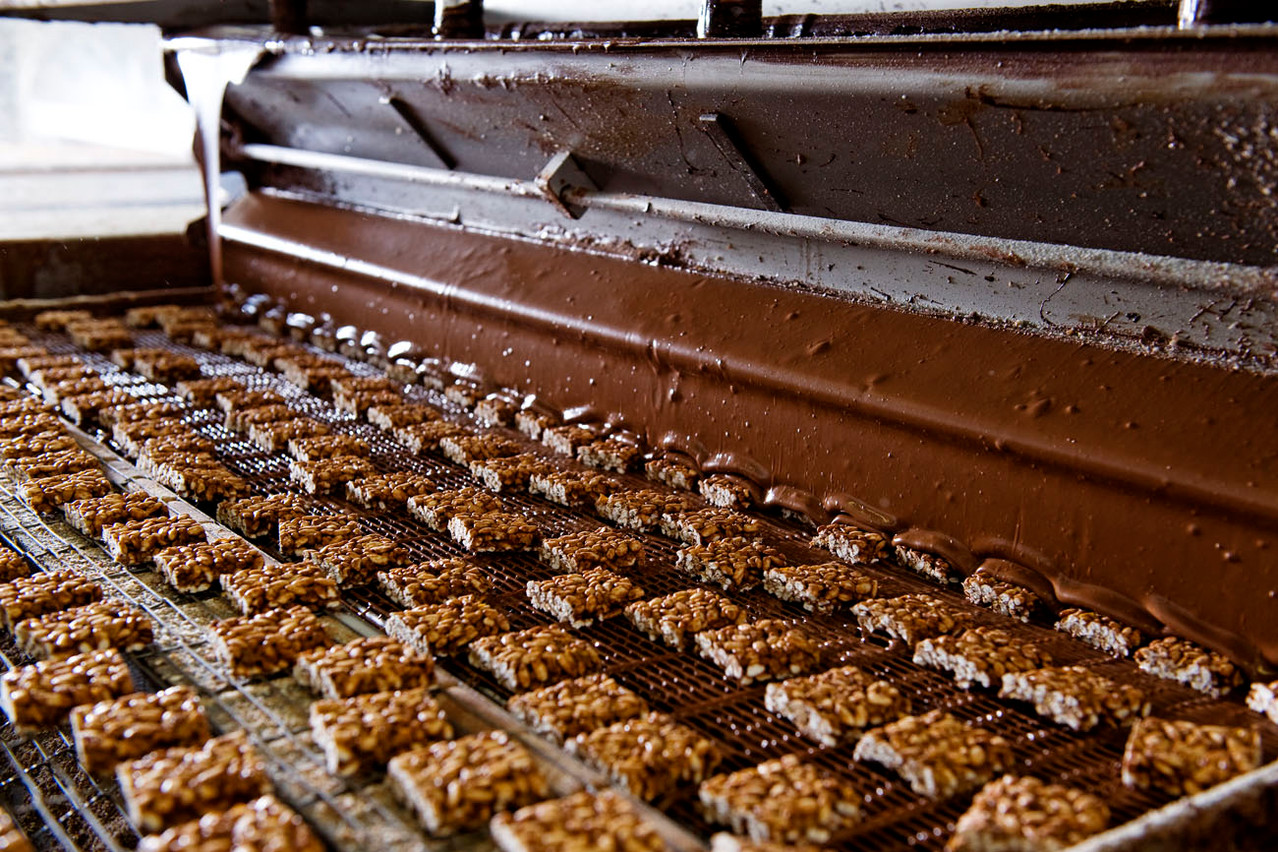 Die Sun Rice-Happen fahren auf einem Gitter unter einem Vorhang aus Vollmilchschokolade hindurch. Überflüssige Schokolade tropft nach unten ab.