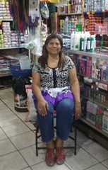 Picture of Claudia's mother sitting in a chair and posing for the camera in front of their El Salvador market.